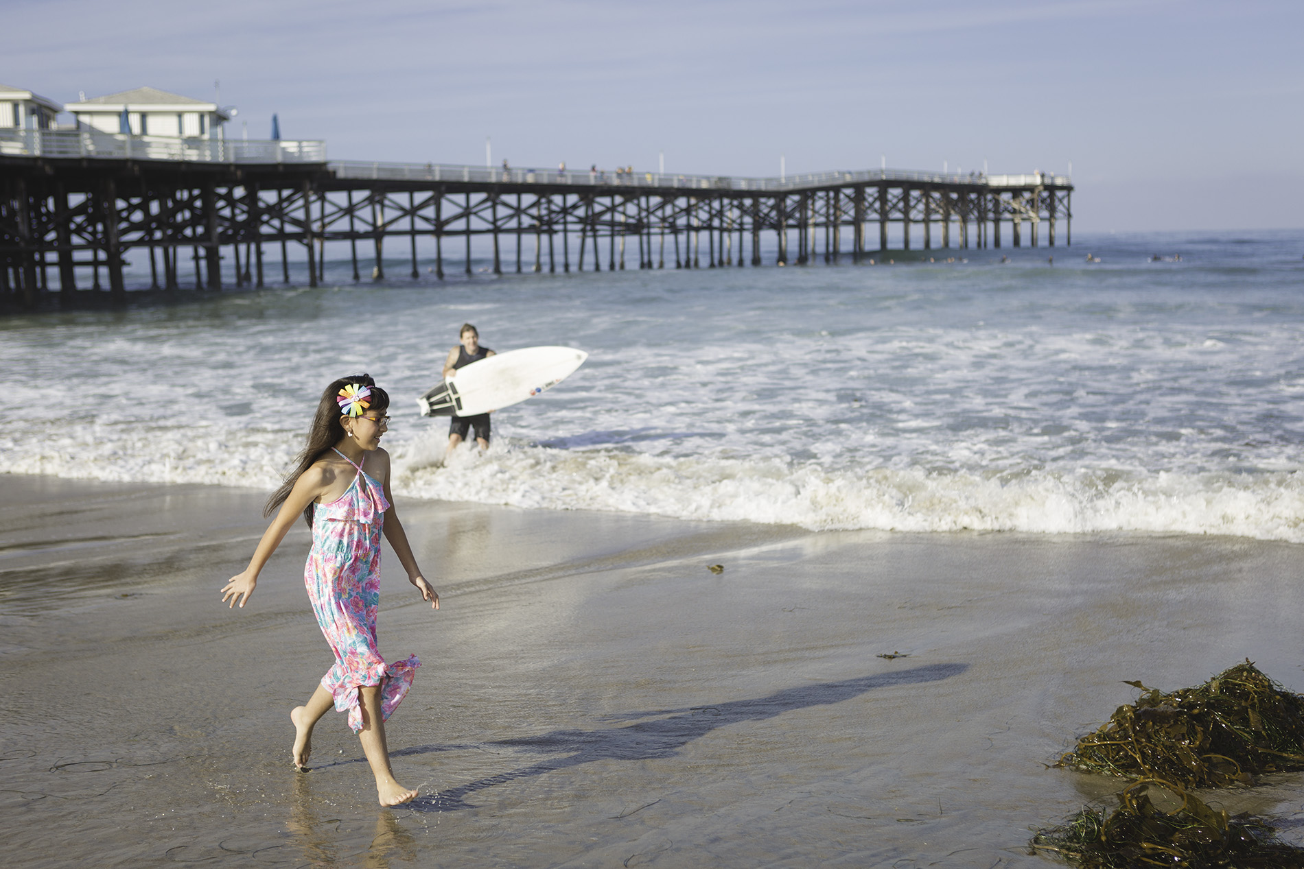 a girl walks on the beach in san diego while on vacation with her family. a surfer is behind her in the ocean.