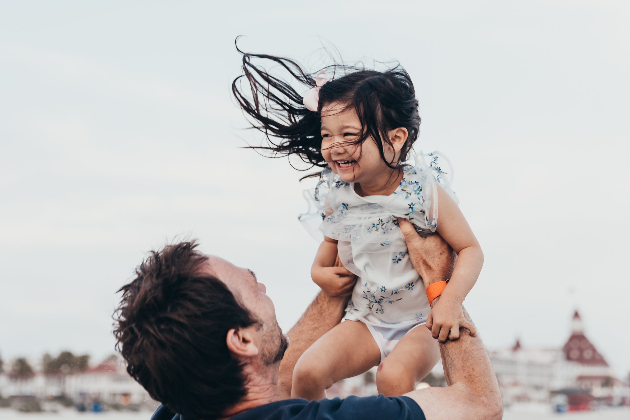 dad plays with daughter at the beach with Hotel del Coronado in the background during their family photography session with Christine Dammann