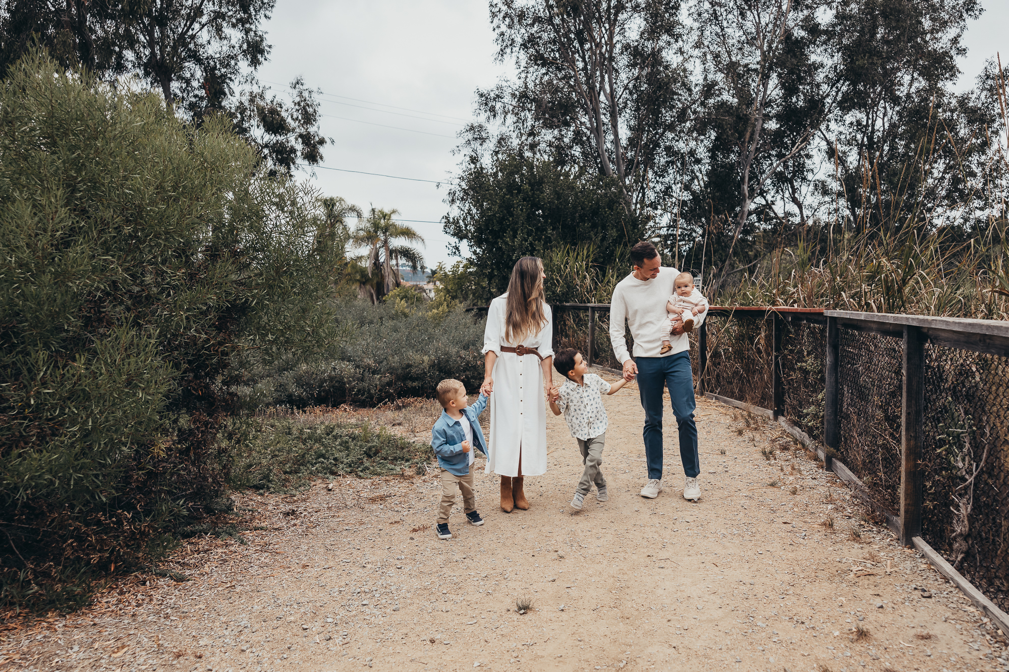 A family of five walks hand in hand on a dirt path surrounded by trees, smiling and enjoying time together outdoors.