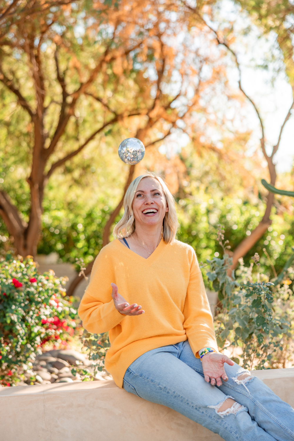 woman in a yellow pullover and light jeans sitting in nature and throwing a small disco ball in the air