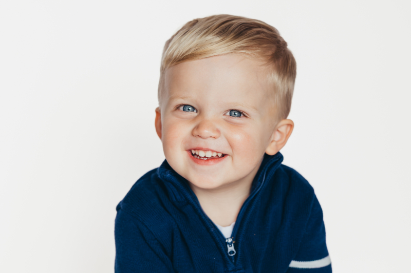 Smiling toddler with bright blue eyes wearing a navy sweater, classic clean studio portrait with soft lighting and simple background.