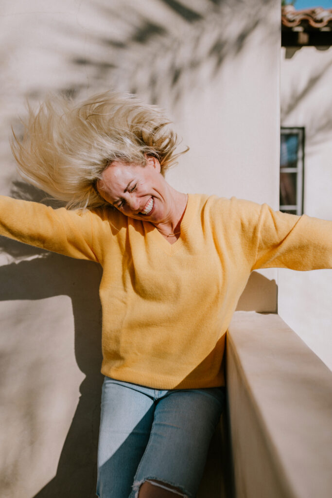 A joyful woman in a bright yellow sweater throws her head back with a big smile, hair flying in the sunlight, against a neutral wall with soft shadows of palm leaves. | photography education