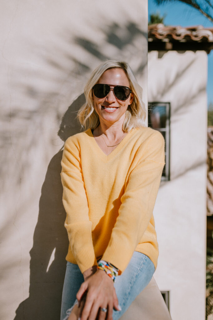 a woman smiling during golden hour with sunglasses on wearing a bright orange sweater 