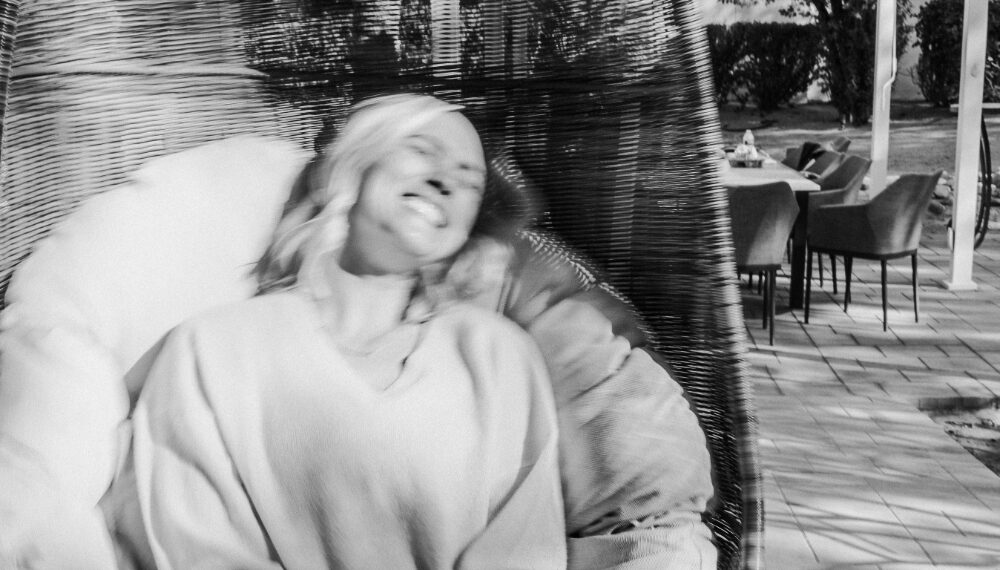 Black-and-white photo of a woman smiling with eyes closed while swinging in a hanging chair outdoors, creating a sense of motion blur and joyful energy.