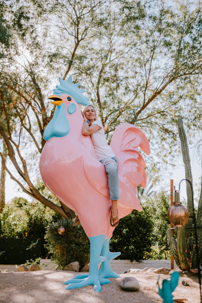 Woman in casual outfit sits playfully on a large pink rooster statue with bright blue details, surrounded by desert plants and tall trees.