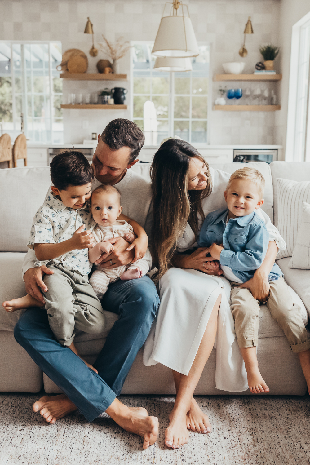 A family cuddles on a white couch in a sunlit living room, sharing a relaxed and joyful moment with their young children and baby | how to share family photos