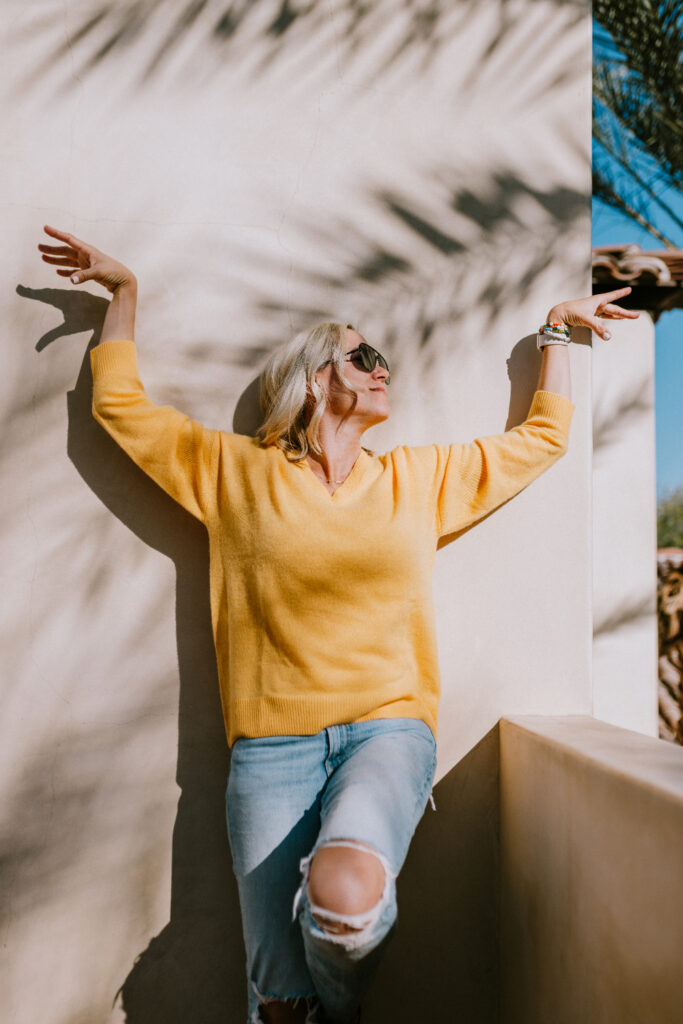 A stylish woman in a yellow sweater and ripped jeans poses against a sunny stucco wall, striking a carefree pose with shadowy palm leaves behind her | how to share family photos