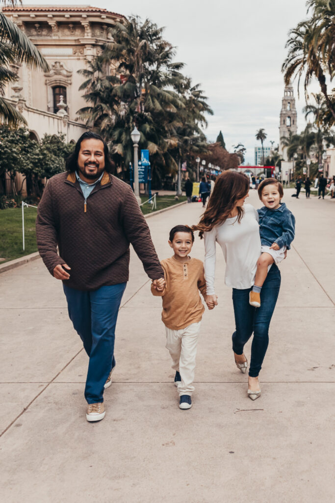 A family of four walks hand-in-hand through Balboa Park in San Diego, smiling and enjoying a candid moment during their photo session.