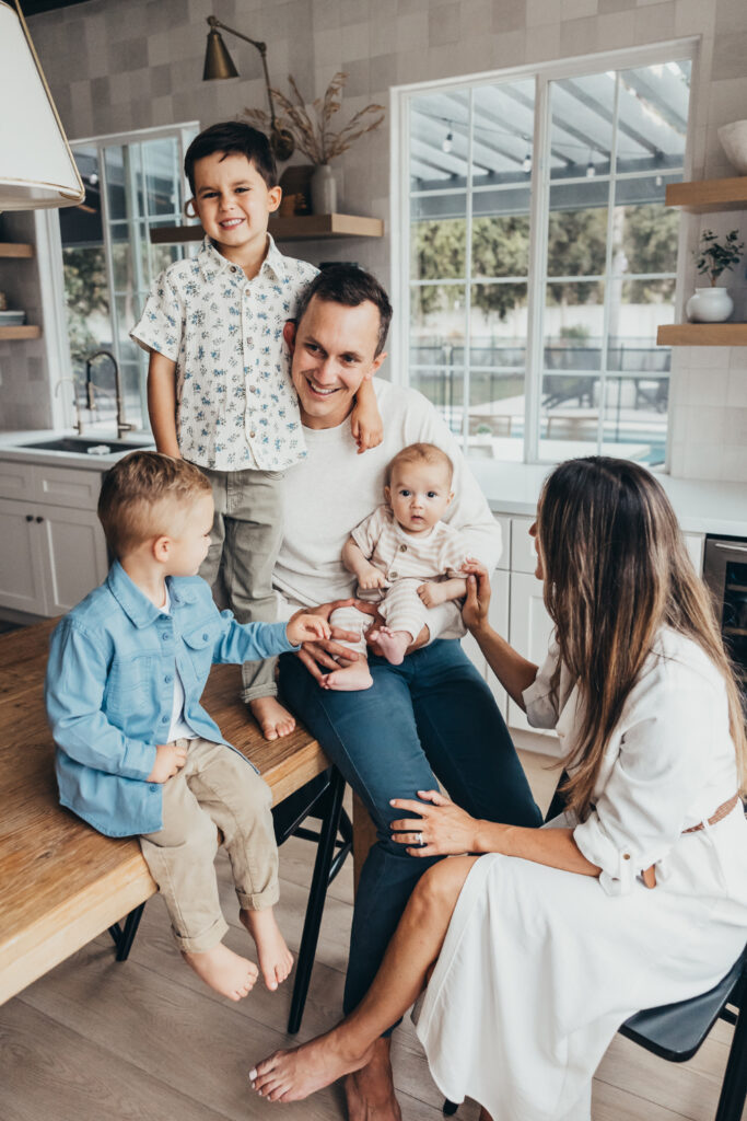 A family of five gathers in a cozy kitchen, with kids sitting on the counter and a baby on dad’s lap—full of natural smiles and sweet sibling interactions | how to share family photos