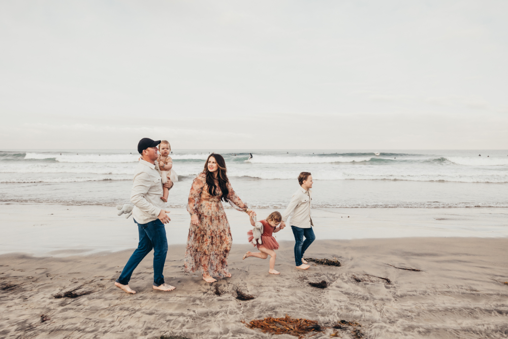 A playful family walks barefoot along the beach during golden hour, with waves crashing in the background and kids running ahead in the sand.