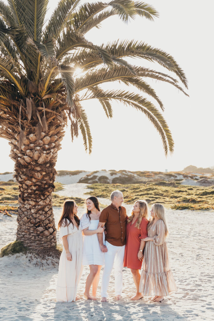 A multi-generational family stands together under a palm tree on the beach in Coronado, laughing and soaking up the sun in coordinated, coastal outfits.