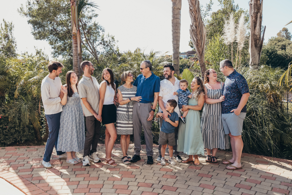 Large extended family standing in a relaxed line in a lush garden, smiling and interacting naturally, capturing a warm, multigenerational group portrait.