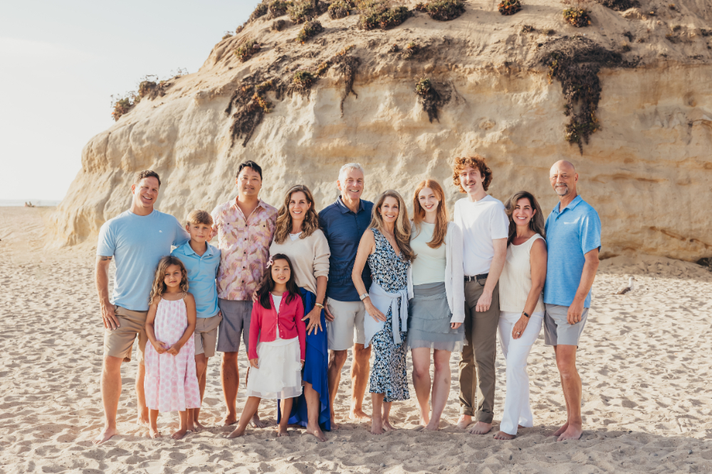 Extended family posed barefoot on a sandy beach with coastal cliffs behind them, arranged in a clean, classic group portrait with a relaxed seaside feel.