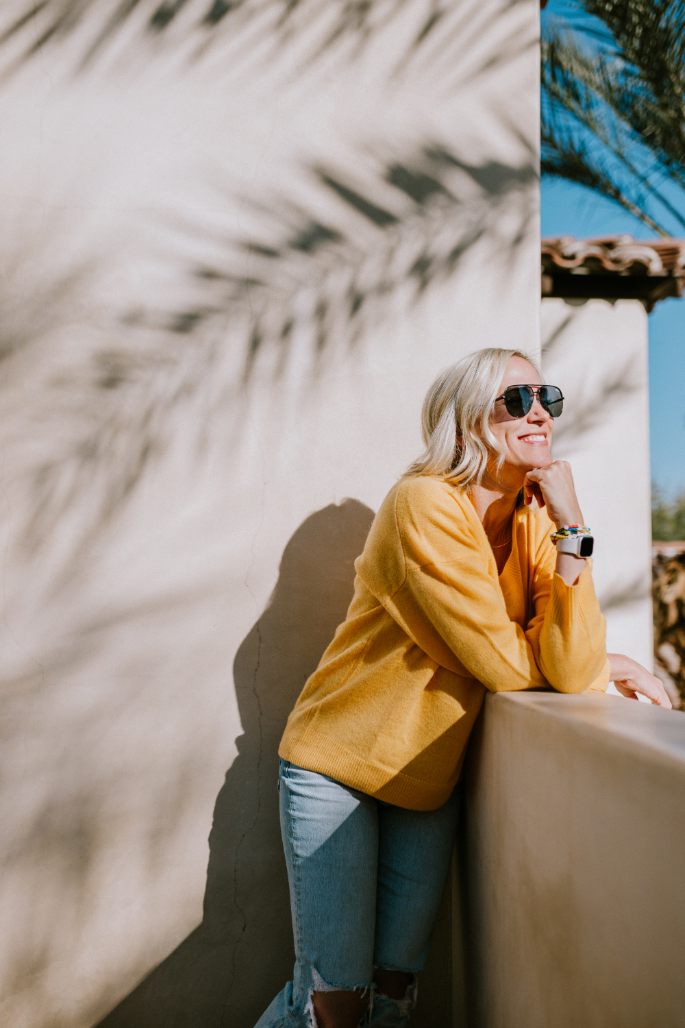 Woman in a mustard sweater and sunglasses leaning on a stucco wall, smiling in warm sunlight with palm shadows creating a calm, lifestyle portrait.