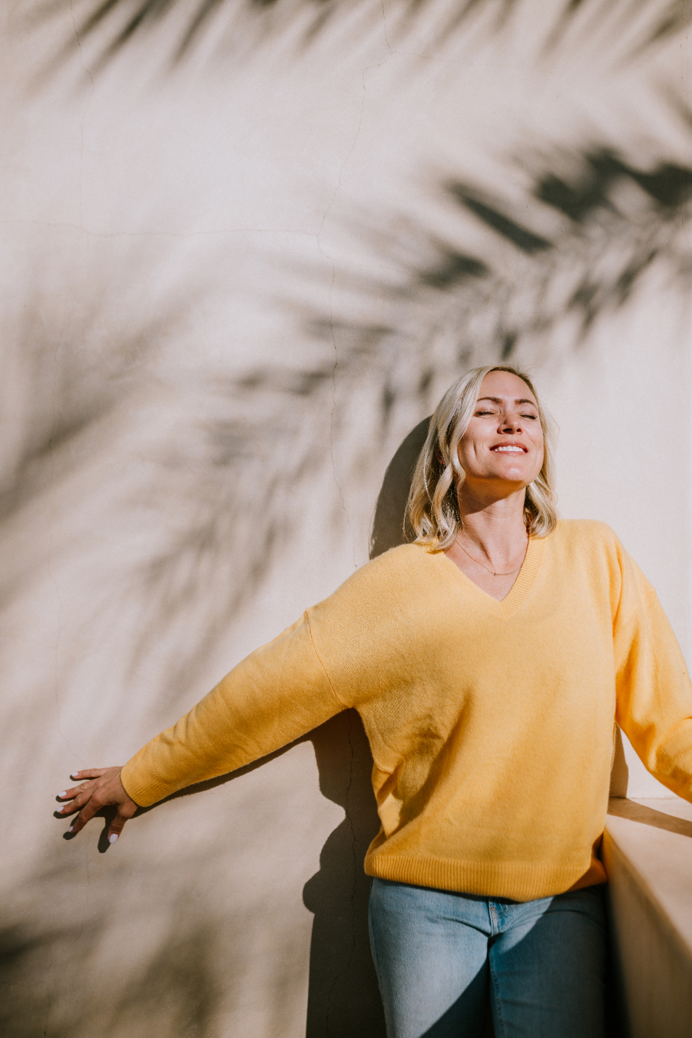 Woman standing against a sunlit stucco wall with arms outstretched, eyes closed, wearing a mustard sweater and jeans, capturing a peaceful, sun-soaked moment.