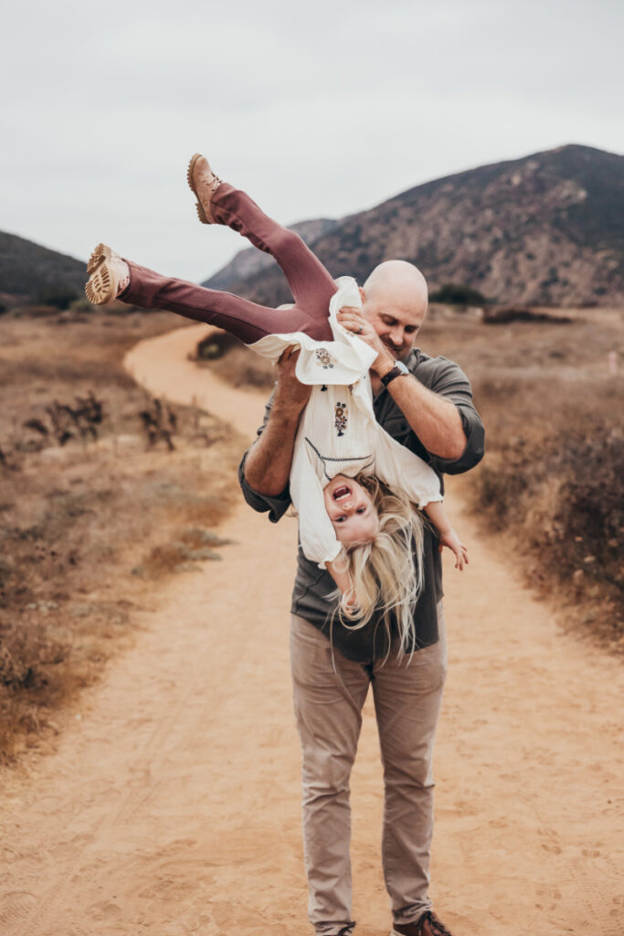 Dad playfully swinging daughter upside down on a dirt path, both laughing during outdoor family session.