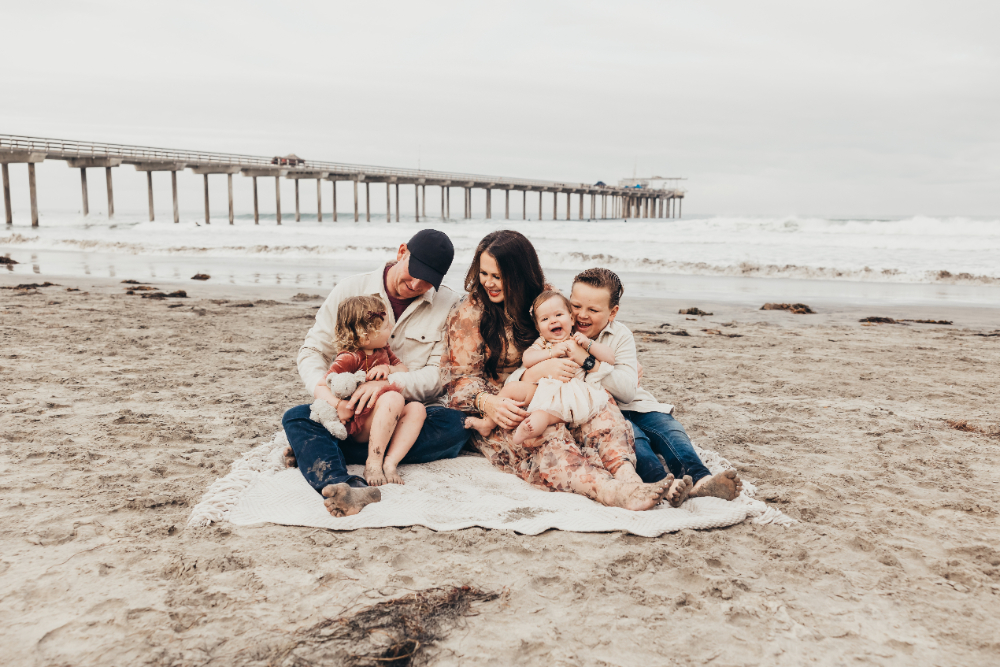 Parents and three young kids cuddling and smiling on the sand during a beach lifestyle photography session.