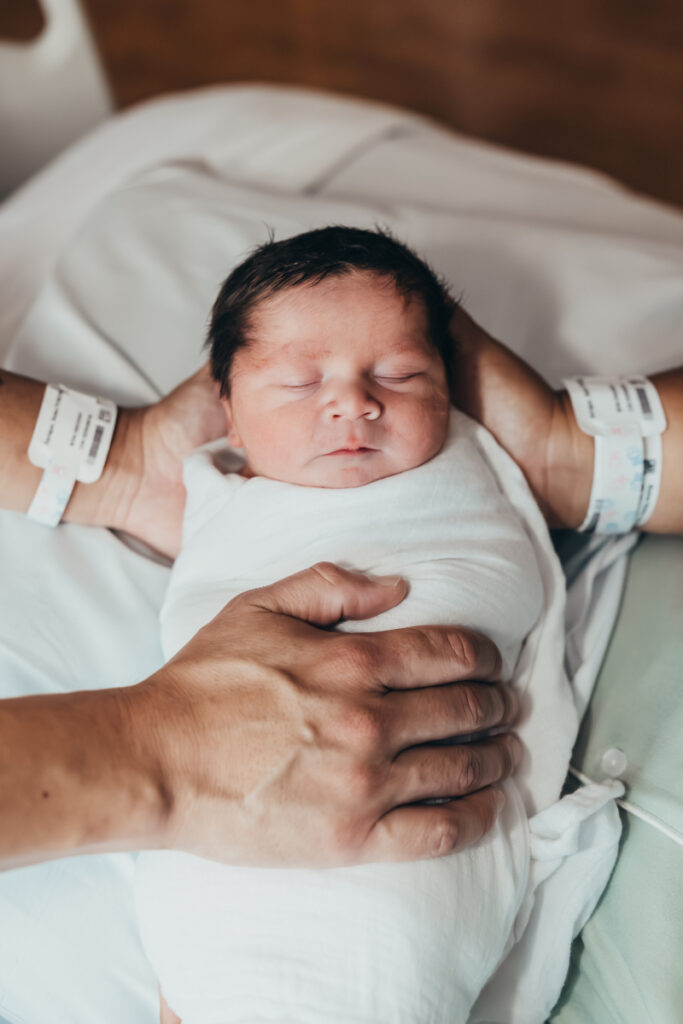 Swaddled newborn resting peacefully in parent's hands, hospital bracelets visible on both parents' wrists.