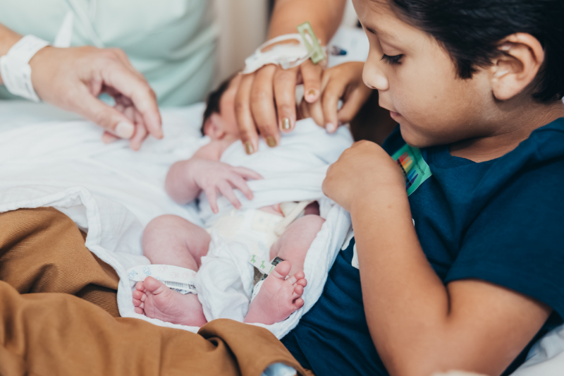 Older sibling meeting newborn during hospital session for newborn photography in San Diego.