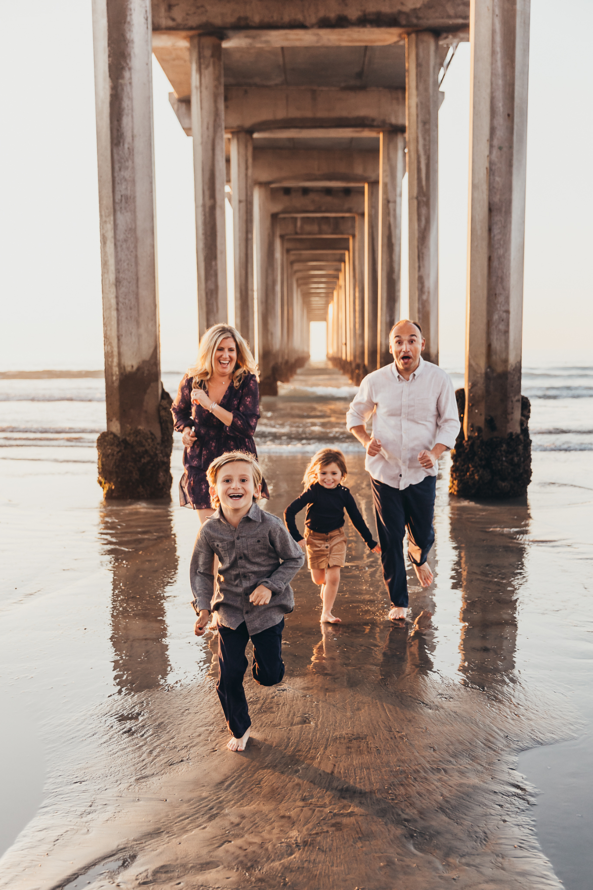 Family of four runs barefoot under a beach pier at sunset, laughing and splashing through wet sand. Joyful candid photos capture real movement and connection in golden light.