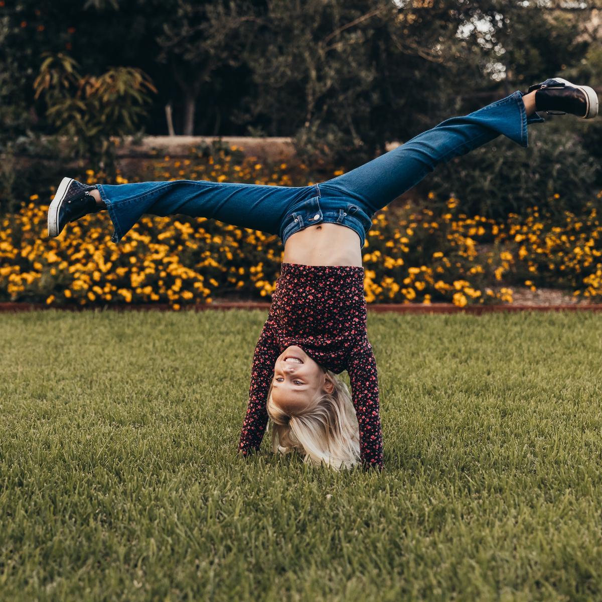 Young girl doing a handstand on green grass in a garden, smiling as her hair falls toward the ground. Playful candid photography moment full of energy and personality.