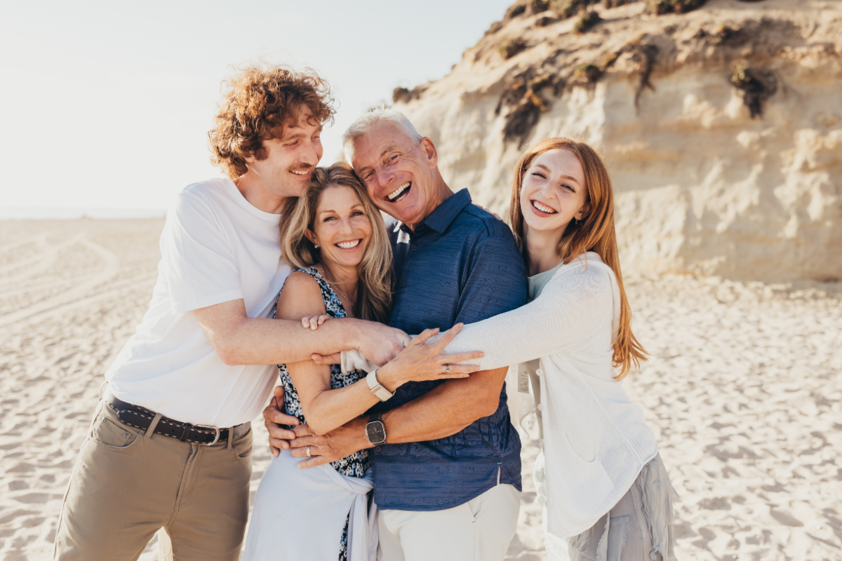 Smiling family of four hugs closely on a sunny beach with sandy cliffs behind them. Warm candid portraits show genuine laughter and relaxed connection by the ocean.