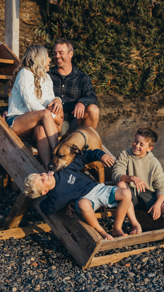 Parents and kids laughing on wooden stairs while dog licks child during relaxed family photo session outdoors.