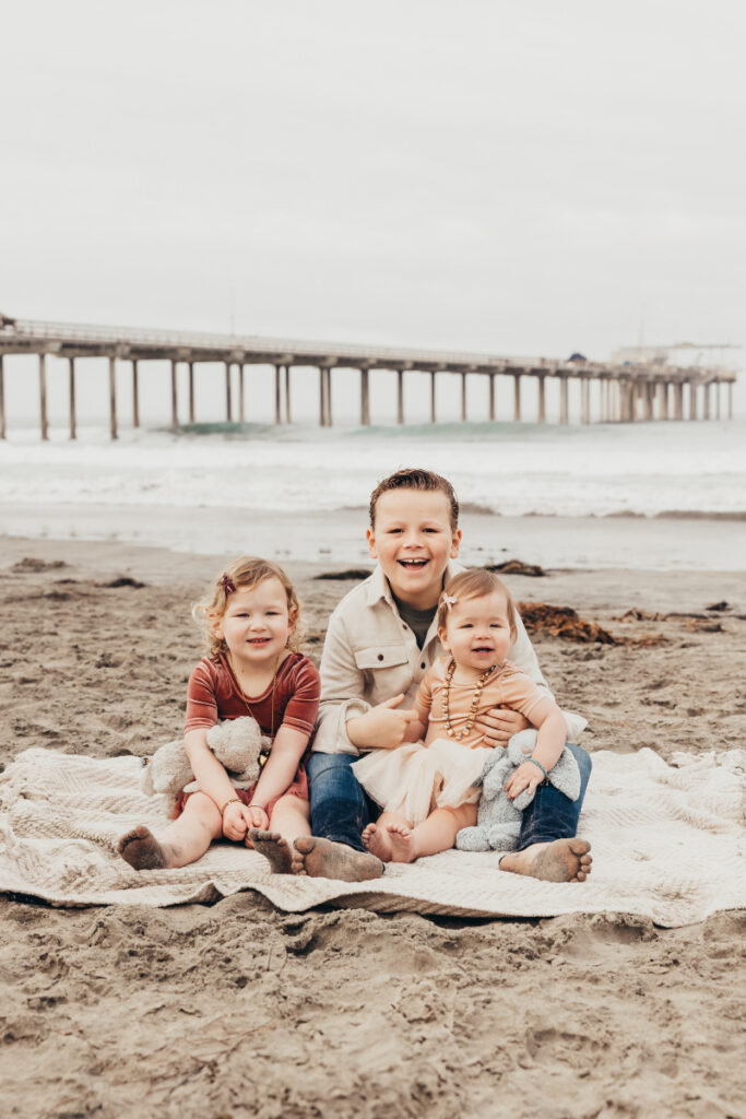 Three kids sitting on a blanket at the beach, smiling with sandy feet and soft ocean background