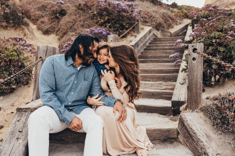Family sitting on wooden beach stairs, parents hugging child, warm candid moment with coastal plants and texture