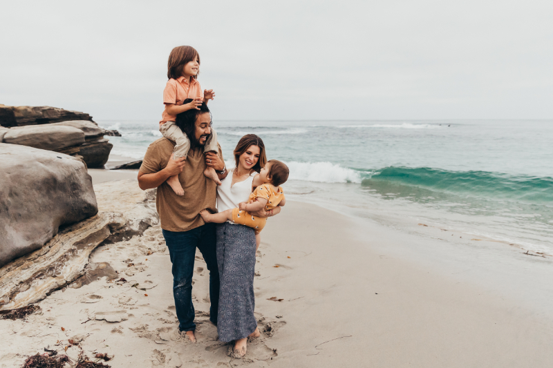 Family standing together near ocean waves, father holding child on shoulders, relaxed beach moment