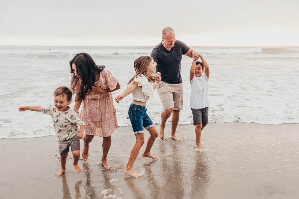 A joyful family runs through the waves at the beach, laughing and holding hands during a candid lifestyle session. Natural moments like this show why connection and emotion matter when learning how to start a photography business that attracts family clients.