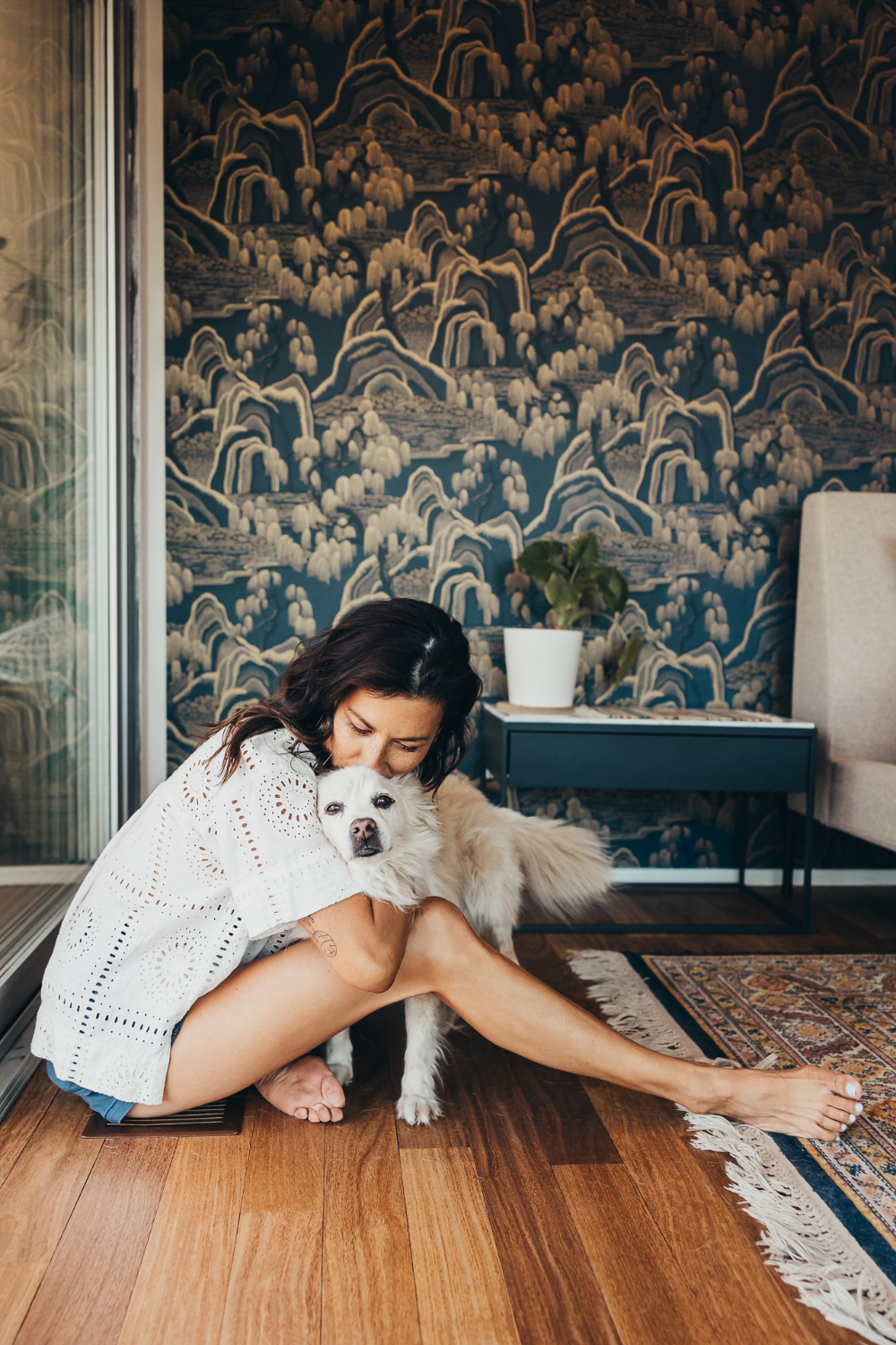 A woman sits indoors hugging her white dog beside dramatic patterned wallpaper and warm natural light. Lifestyle portraits like this help photographers create emotional storytelling images while starting a photography business with a unique personal style.