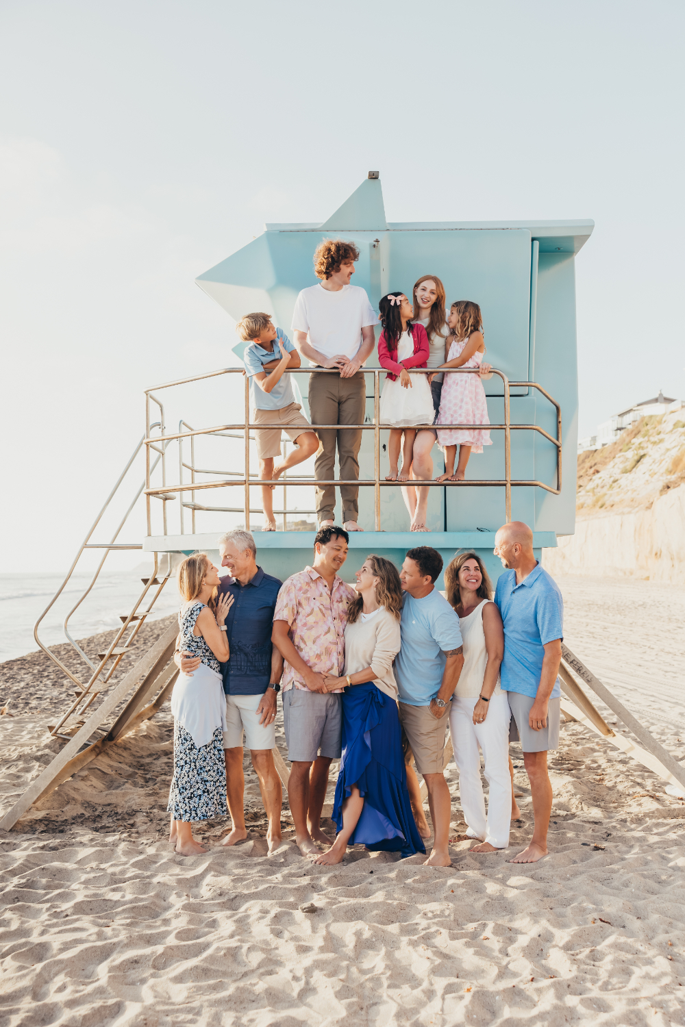 A large multigenerational family poses together beside a pastel blue lifeguard tower on the beach at sunset. Group sessions like this highlight the importance of communication, posing, and client experience when starting a photography business.
