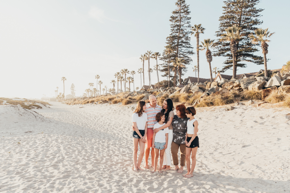 A family gathers together on a wide sandy beach lined with palm trees and coastal homes, sharing a candid moment during golden hour. This natural family portrait highlights connection, laughter, and the timeless beauty of a beach photography session.