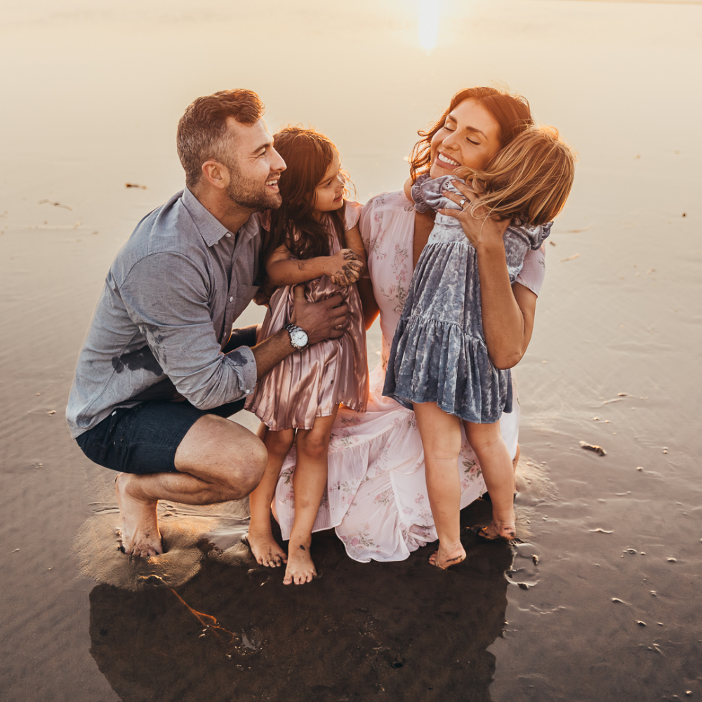 A family of four embraces on the beach at sunset, with parents holding their young daughters close as golden light reflects off the wet sand. This heartfelt family portrait showcases genuine emotion and the meaningful moments that inspire trust within an email funnel strategy.