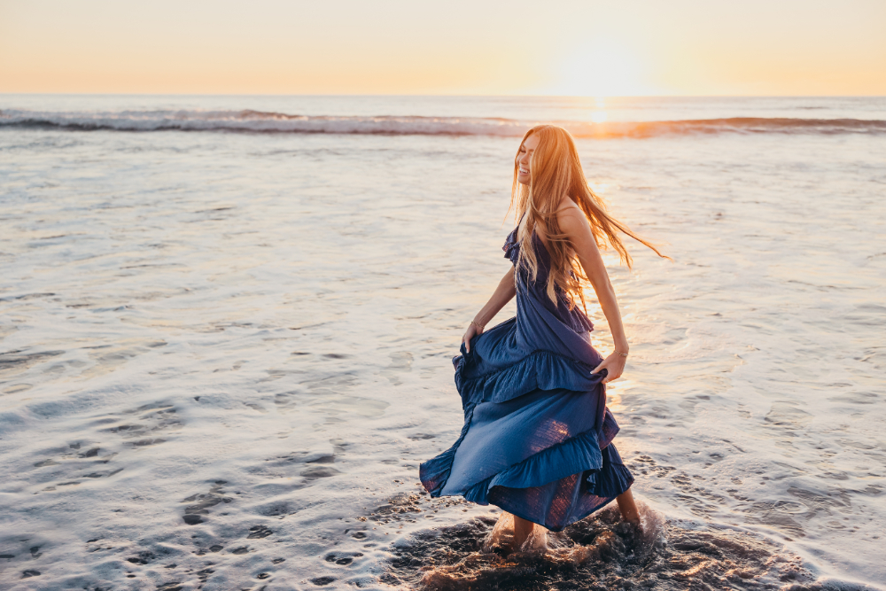 A woman in a flowing blue dress walks through shallow ocean water at sunset, smiling as golden light reflects across the waves. This dreamy beach portrait captures movement, warmth, and the carefree beauty of a coastal photography session.