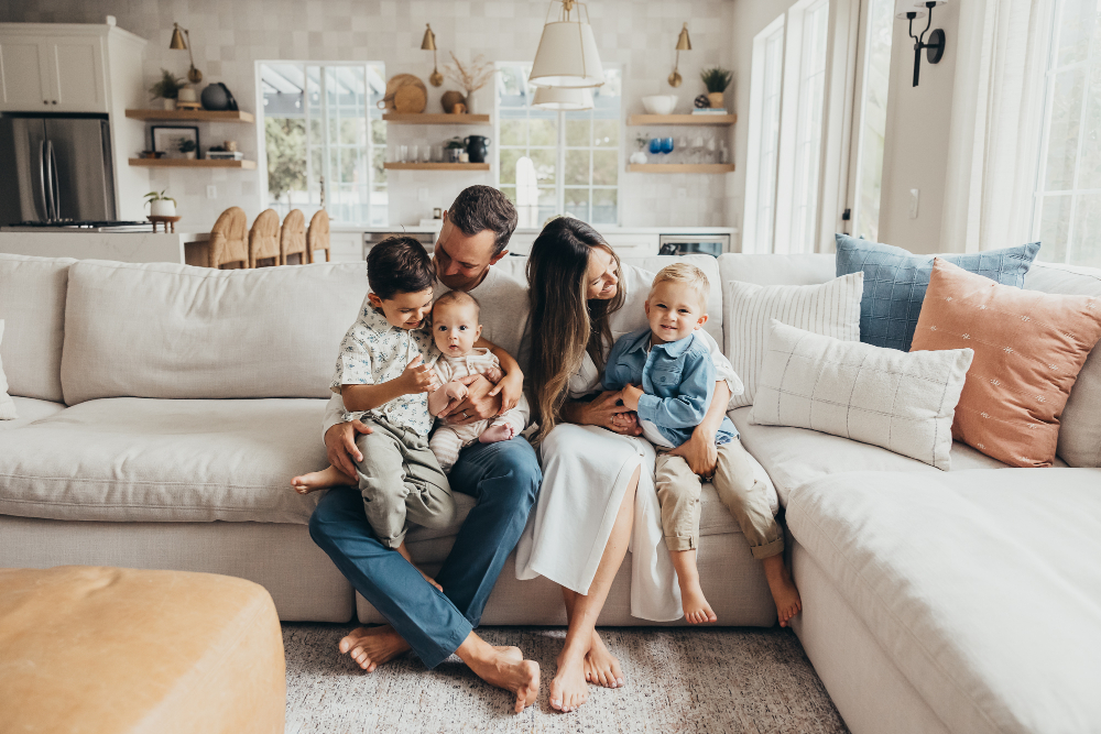 A family of five cuddles together on a bright living room sofa, sharing smiles and sweet moments with their children. This lifestyle family portrait showcases genuine connection and helps illustrate what clients can expect when exploring photography pricing.