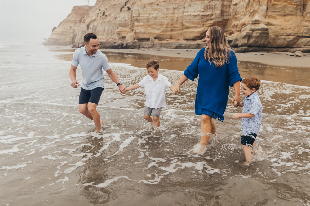 Parents and their two young sons splash through the shoreline while holding hands beneath dramatic coastal cliffs. This candid beach family session highlights playful moments, authentic emotions, and the value behind photography pricing for lasting memories.