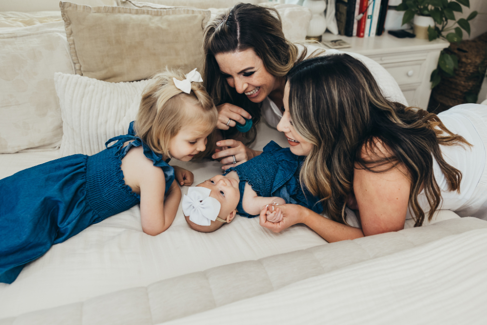 Two mothers and their young daughters share a tender moment on a cozy couch, smiling and engaging with a newborn baby. This in-home lifestyle portrait celebrates family bonds, love, and the beauty of everyday moments together.