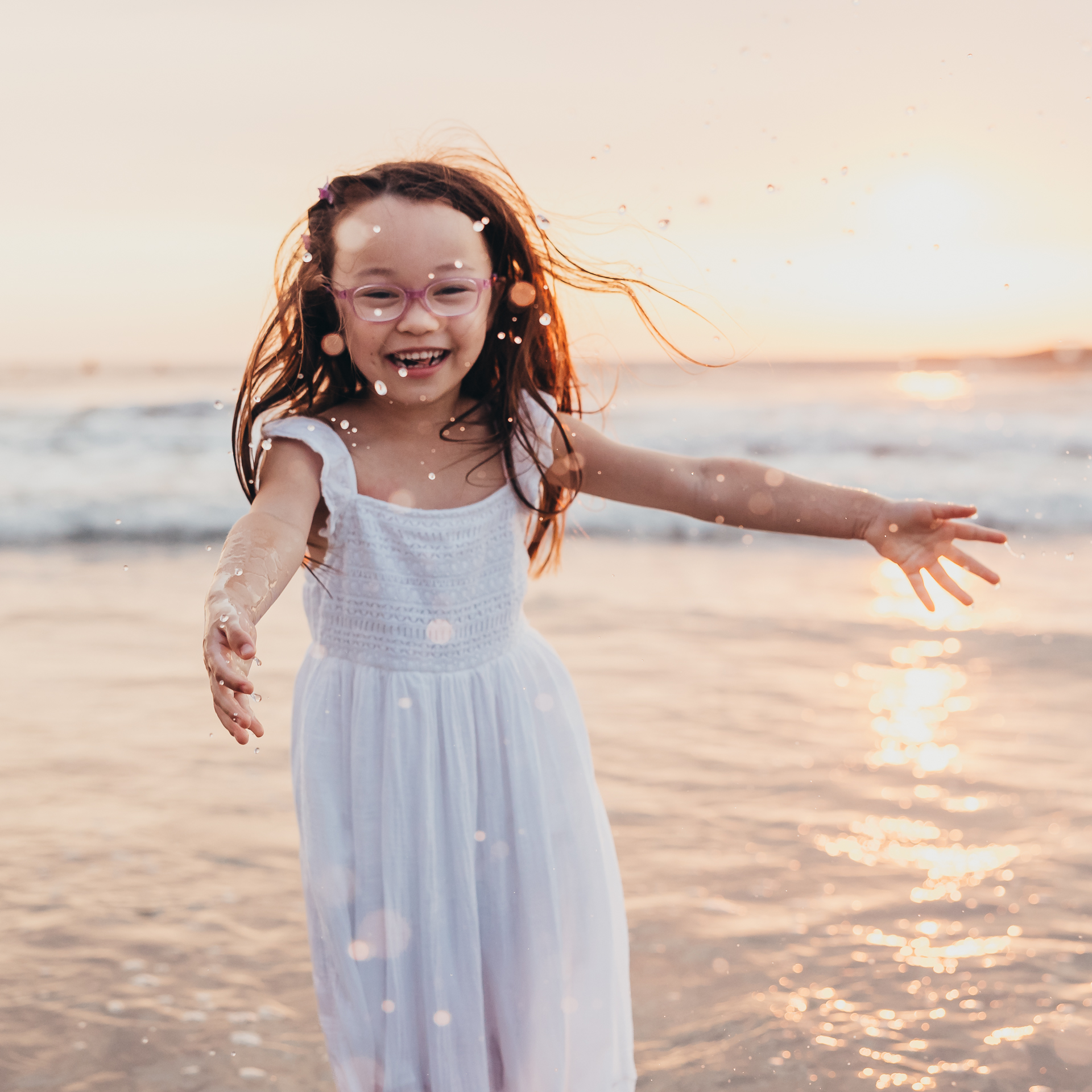 A joyful young girl runs toward the camera at the beach during golden hour, smiling as ocean spray sparkles around her. This playful moment beautifully captures the heart of preserving memories through genuine laughter, movement, and carefree childhood joy.