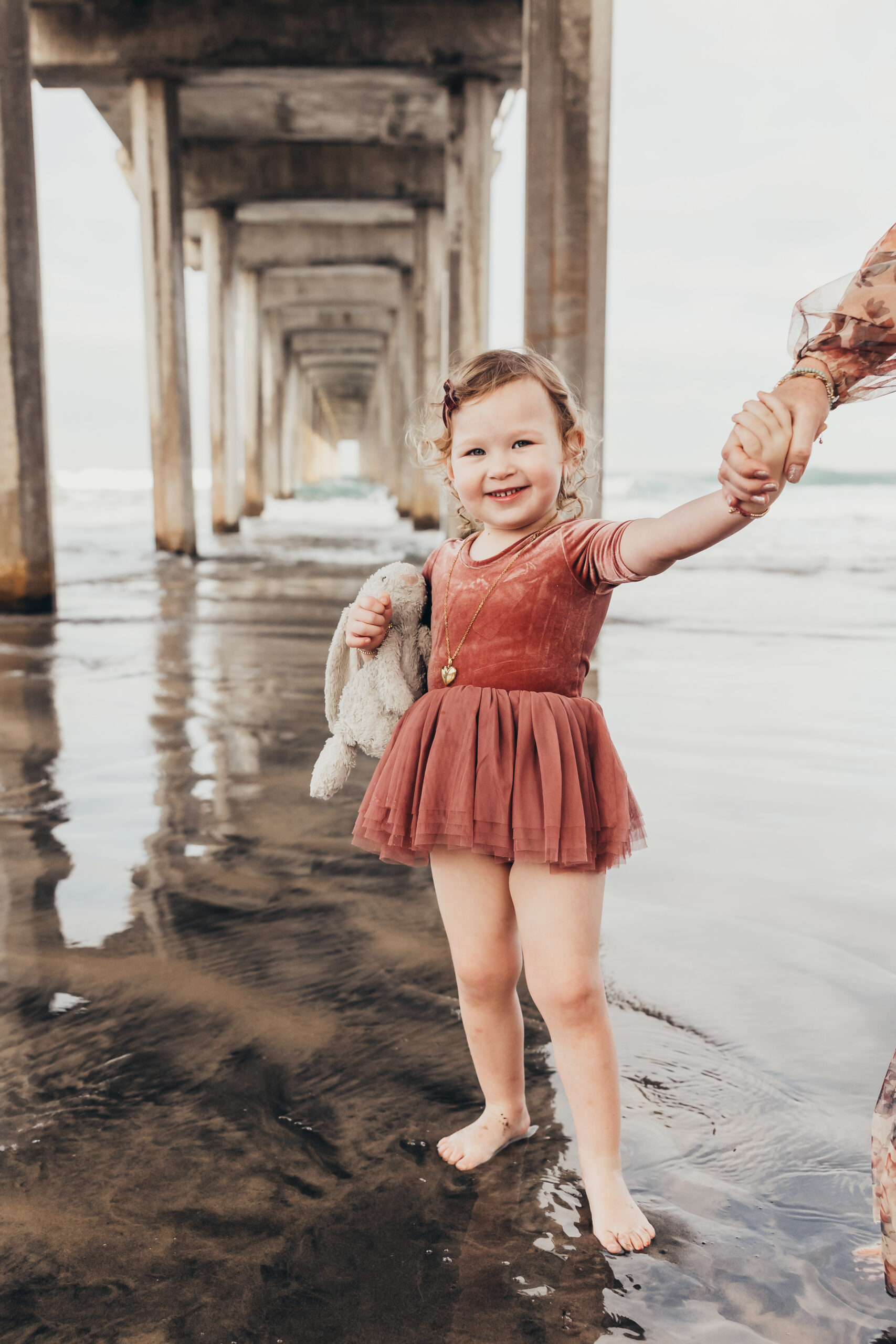 A smiling toddler walks barefoot beneath a beach pier while holding a favorite stuffed bunny and reaching for a parent's hand. This sweet, candid portrait highlights the beauty of preserving memories through everyday childhood moments and treasured comforts.