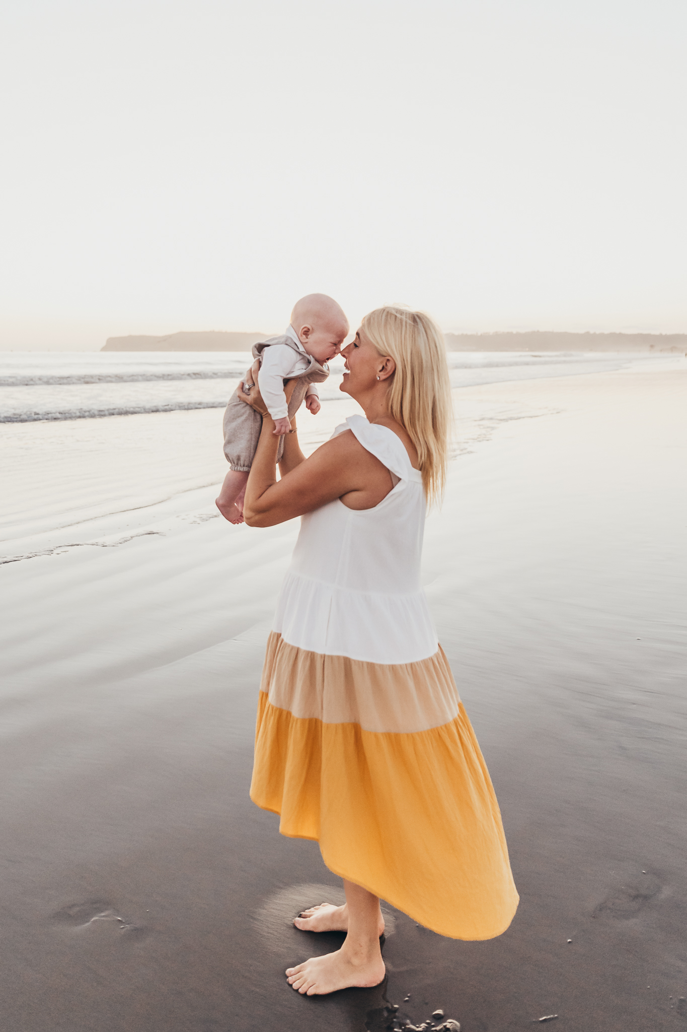 A mother lifts her smiling baby into the air on a peaceful beach at sunset, sharing a tender nose-to-nose moment. The soft light and genuine connection create a timeless family portrait celebrating love, joy, and the fleeting beauty of early parenthood.
