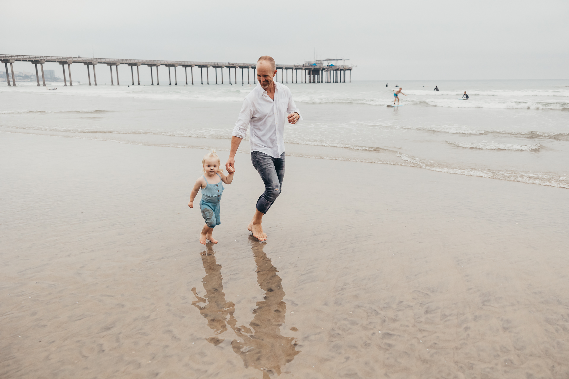 A father walks hand in hand with his toddler along the shoreline near a pier while gentle waves roll in behind them. Their relaxed stroll and shared smiles capture the simple, meaningful connection that makes everyday family moments so special.