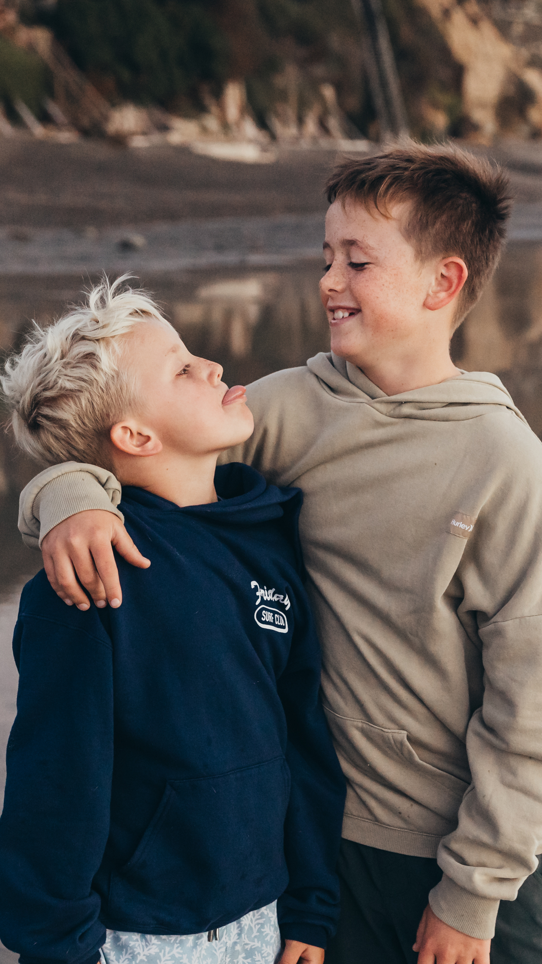 Two brothers stand arm in arm at the beach, laughing together as one playfully sticks out his tongue. Their genuine smiles, sibling bond, and relaxed personalities create a heartfelt portrait that celebrates the fun, connection, and joy of growing up together.