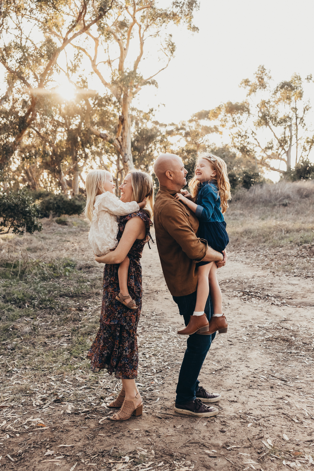 A family of four shares a playful moment on a wooded trail at golden hour, with each parent holding a young daughter. Building genuine family connections and a relaxed experience can be an important part of getting more clients through repeat sessions.
