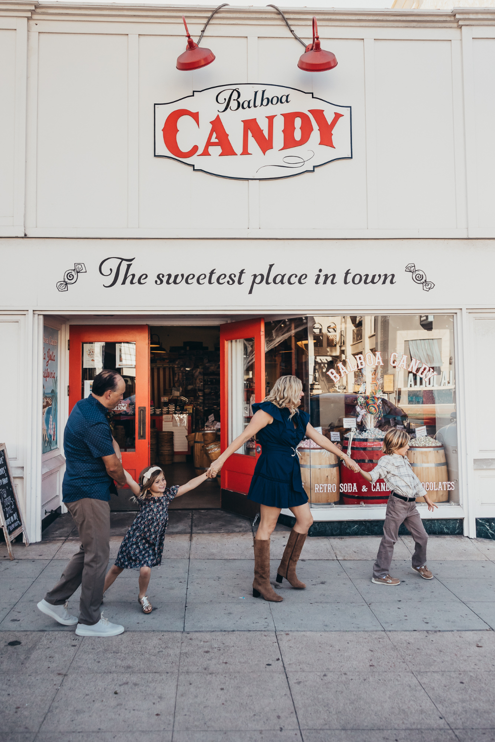 A family of four walks and plays together outside Balboa Candy, with the parents holding their daughter’s hands as their son runs beside them. Choosing fun, unexpected locations can make repeat family photography sessions feel fresh and personal.