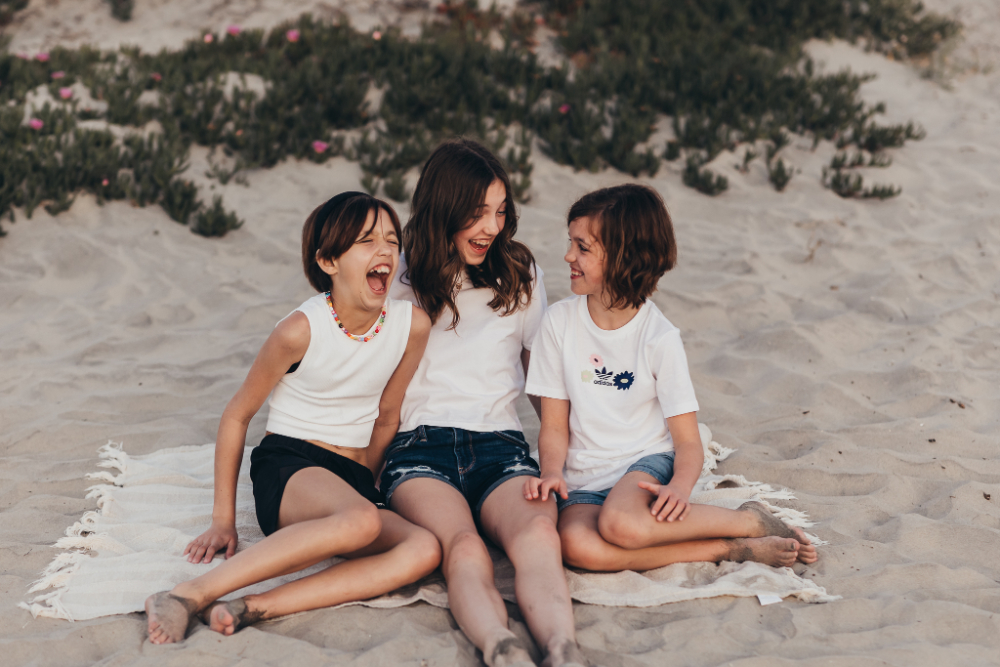 Three siblings sit together on a blanket at the beach, laughing and looking at each other as the sun begins to set. Their joyful expressions and playful connection create the kind of natural family photos parents treasure for years.