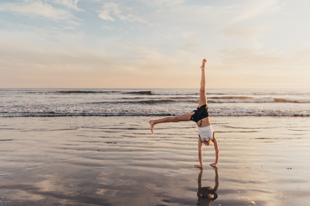 A child performs a cartwheel along the shoreline during sunset, with gentle waves and colorful skies in the background. The playful movement and freedom perfectly capture childhood energy and personality during a family photography session.