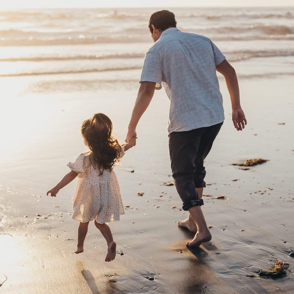 A father and his young daughter walk hand in hand along the shoreline as the sun sets over the ocean. Their quiet connection and carefree steps create beautiful natural family photos filled with warmth, movement, and genuine emotion.