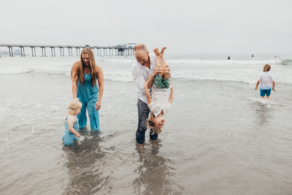 A family splashes in the shallow ocean while a father playfully holds his child upside down and siblings explore nearby. The laughter, movement, and real interactions make these natural family photos full of personality and fun.